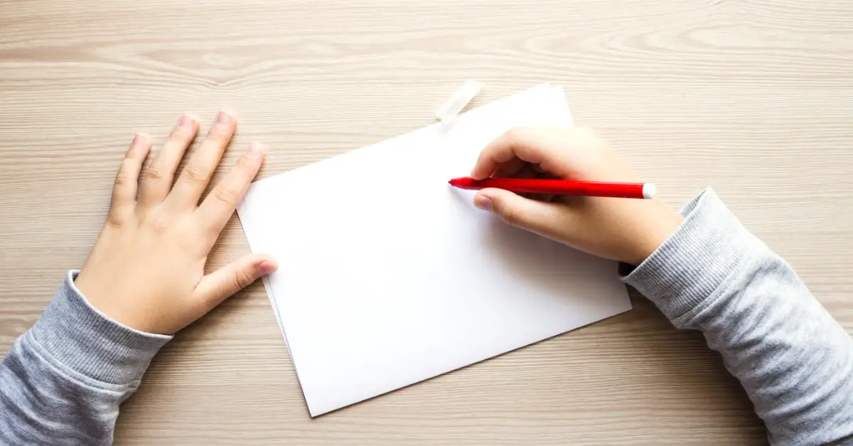 child holding red pen writing on a white piece of paper