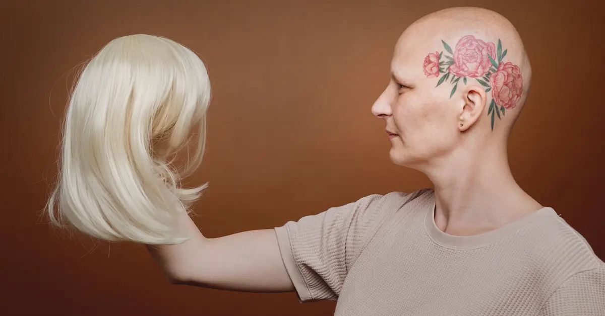 Side view portrait of confident bald woman holding wig of blonde hair against brown background in studio, alopecia and cancer awareness