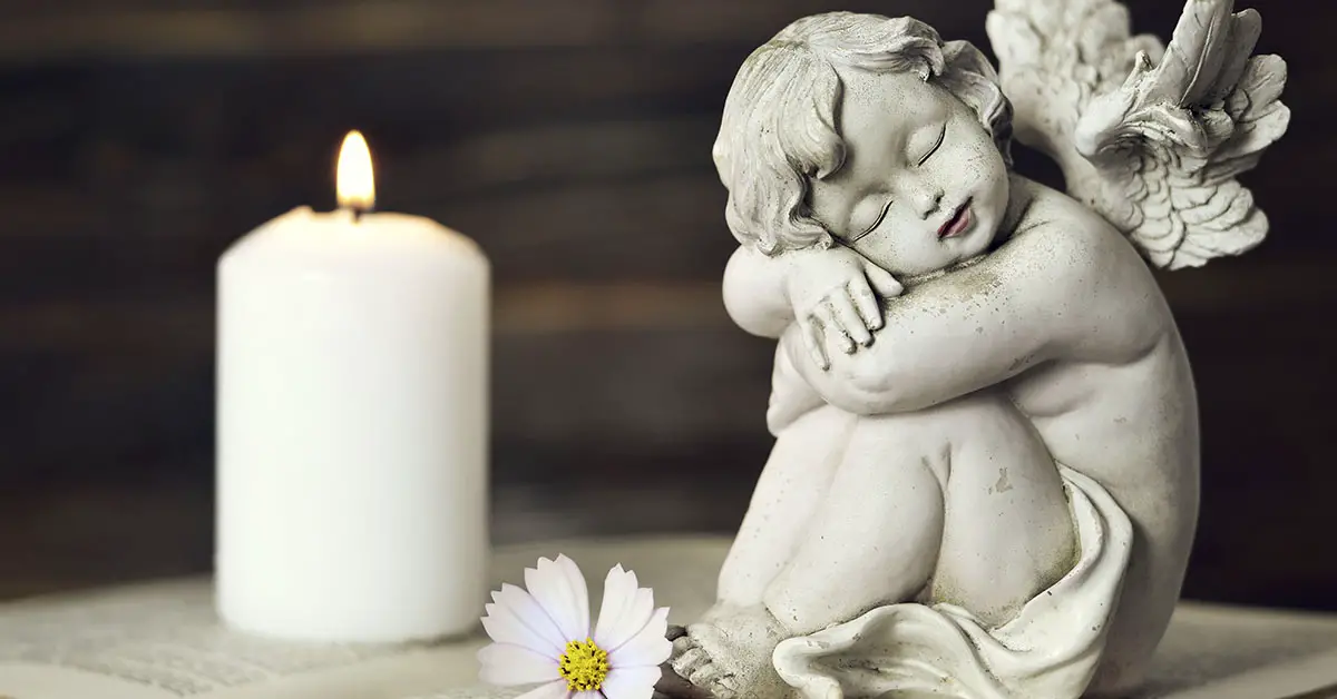 sculpture of an angel sitting bside a lit white candle and a single daisy lays at its base