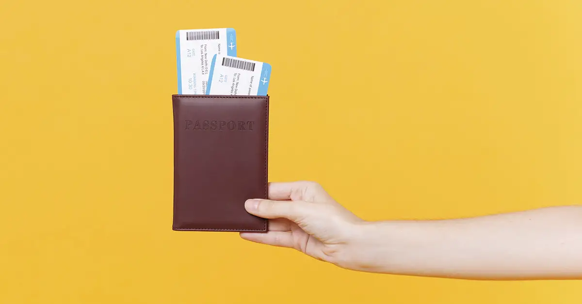 yellow background, arm extended holding brown leather passport holder with two plane tickets coming out of the top