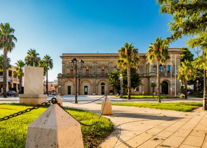 Acquarica del Capo, Presicce, Salento, Puglia, Italy. Town square with the seat of the municipality and the town hall. The ancient stone and brick building. Flower beds with the lawn and palm trees.