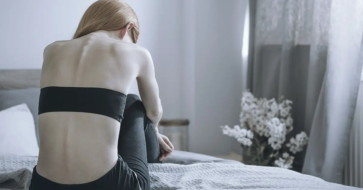 underweight woman sitting on bed, upright in fetal position