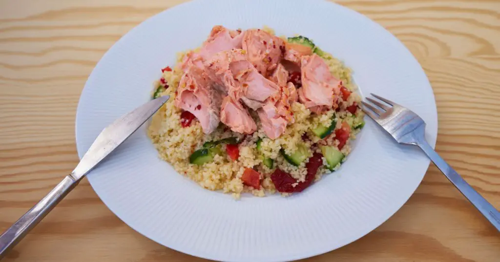 Close up fresh, tasty and healty couscous salad with poached salmon served on simple white plate with fork and kife like light diner or lunch ideal for reduction diet.
