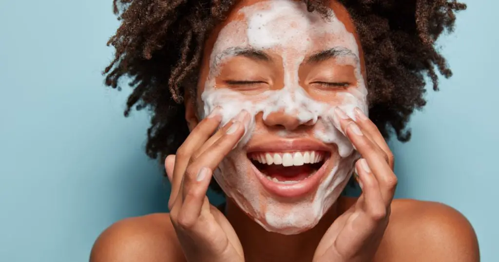 Spa, therapy and beauty concept. Satisfied black woman washes face with white soap, has cleansing foam, purification of face, cleans pores, shows bare shoulders, isolated over blue studio wall
