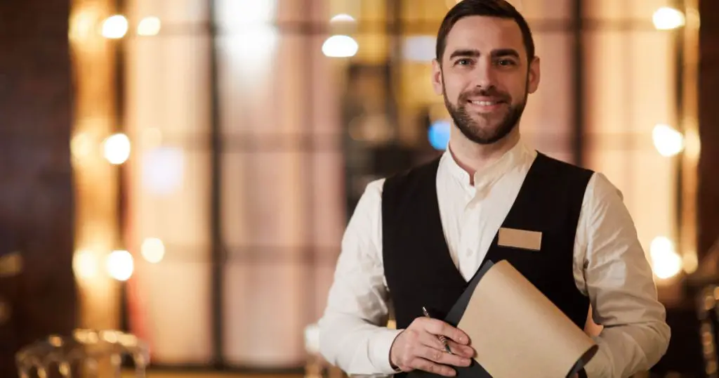 Waist up portrait of handsome waiter smiling cheerfully at camera standing in luxury restaurant or cafe, copy space
