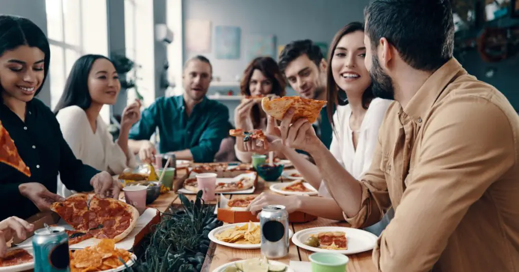 Holiday among friends. Group of young people in casual wear eating pizza and smiling while having a dinner party indoors
