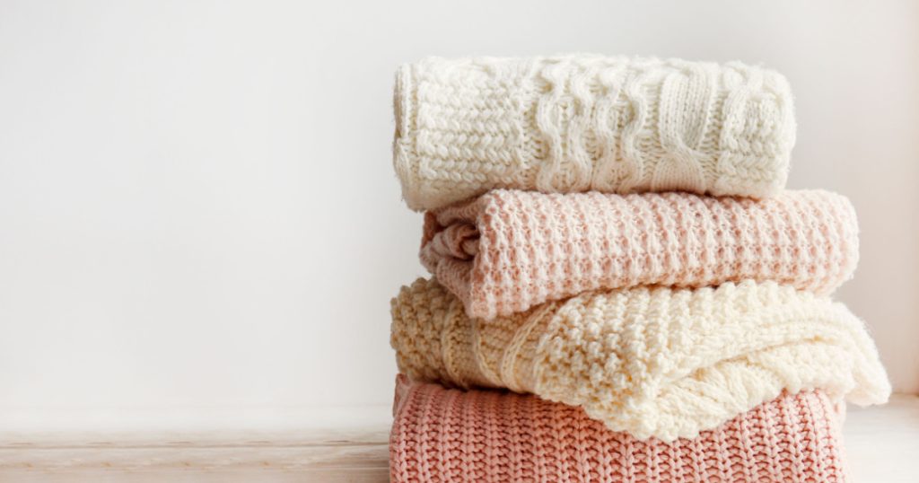 Stack of clean freshly laundered, neatly folded women's clothes on wooden table. Pile of shirts and sweaters on the table, white wall background. Copy space, close up, top view.
