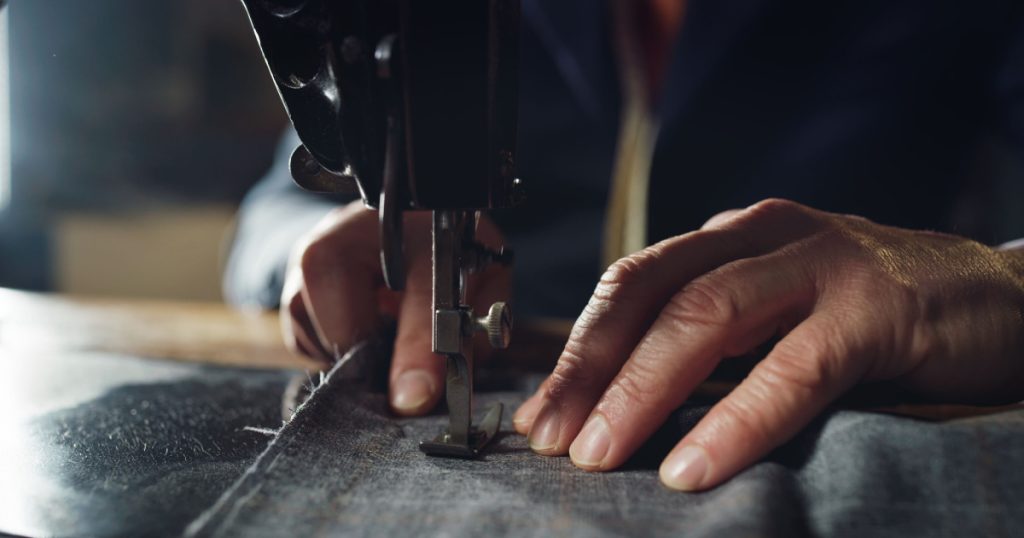 Macro shot of experienced tailor is sewing custom handmade high quality apparel in ancient luxury traditional tailoring workshop. Concept of industry, handmade, hand craft, couturier and tradition.
