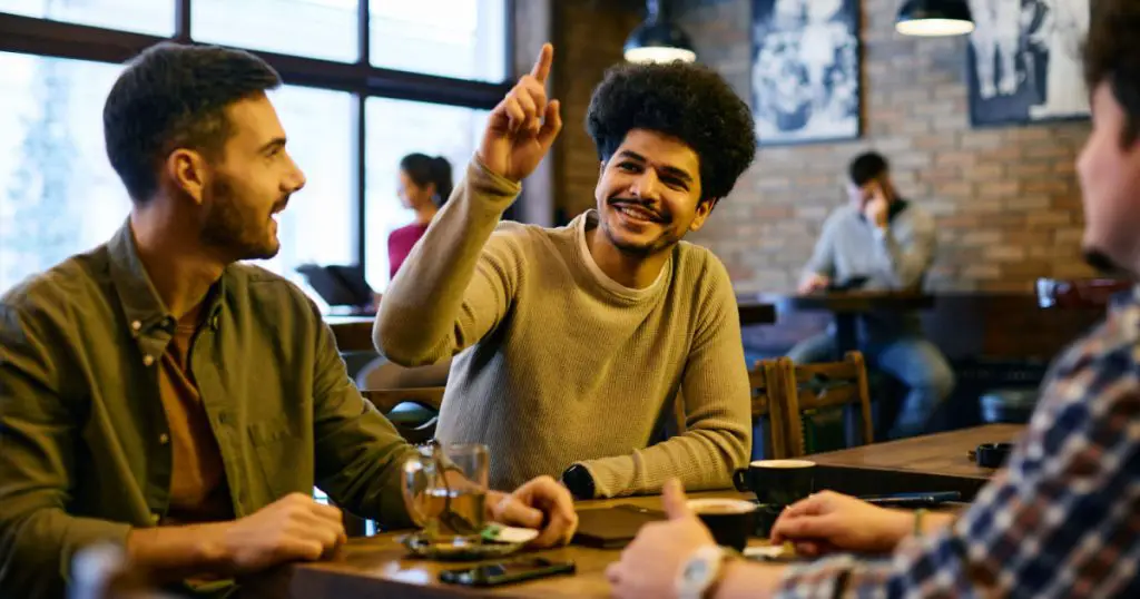 Group of multiracial friends making order in a pub. Focus is on Lebanese man with hand raised calling the waiter.
