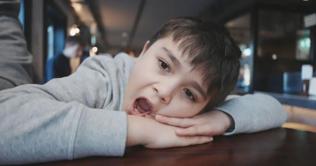 Kid looking out with bored face, Indoors Portrait preschool child laying head down on hand and yawning, Young boy sitting in Resturant or Cafe waiting for food
