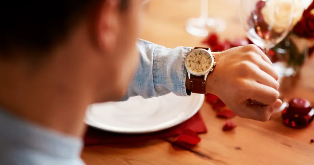 Man checking watch, time on date at dinner table and waiting for person at a restaurant reservation. Impatient guy alone at coffee shop, stress of modern dating and closeup of looking at wristwatch
