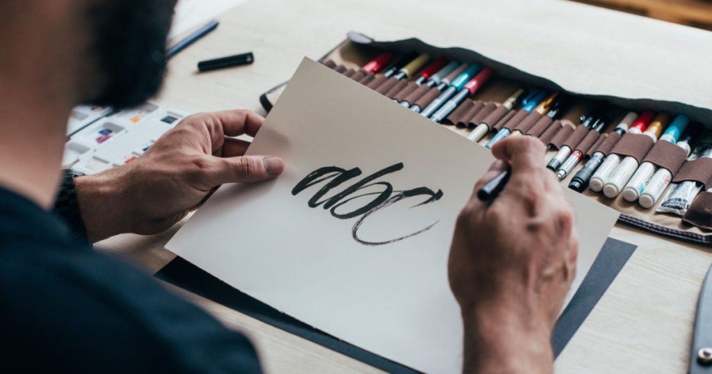 Calligraphy artist or student practices spelling words and letter with black ink brush on white canvas, sits behind table with different kinds of tools, pens, brushes and watercolors set up
