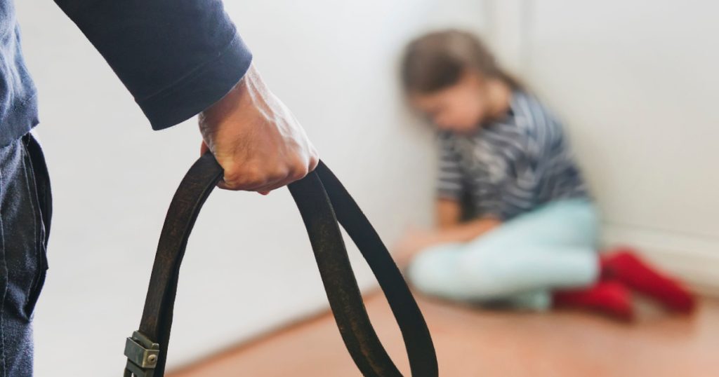 The girl is sitting in the corner hiding from her abusive stepfather. The concept of raising children through domestic violence. Punishment for a crime with a belt. The problem of child abuse.
