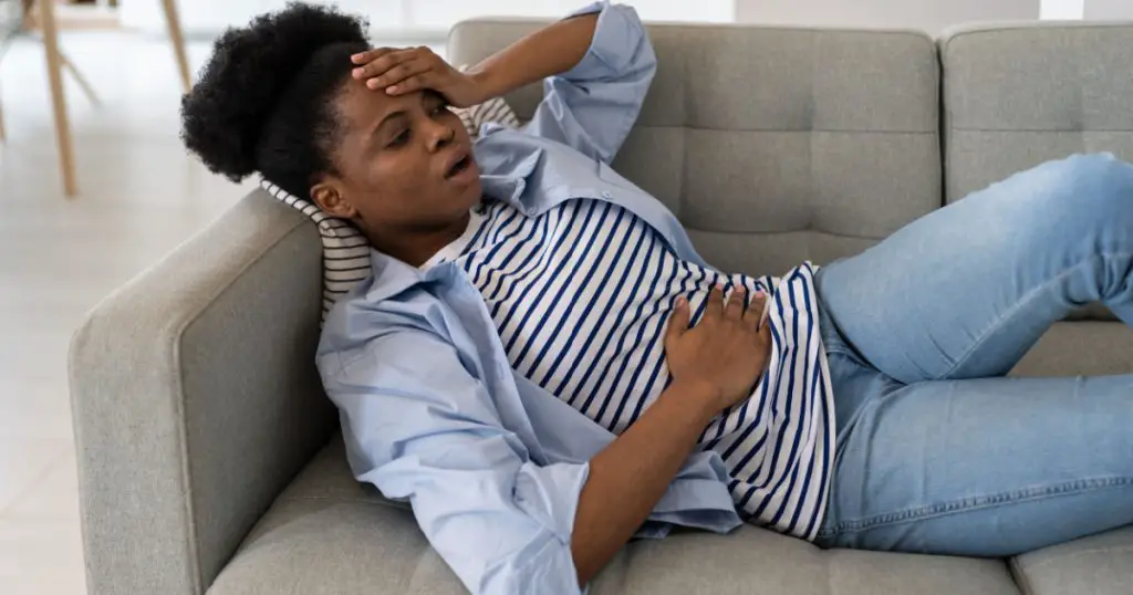 Sick young African American woman having health problems holding head and belly during menstruation. Black girl lying in living room on couch suffering from pain associated with gynecological causes
