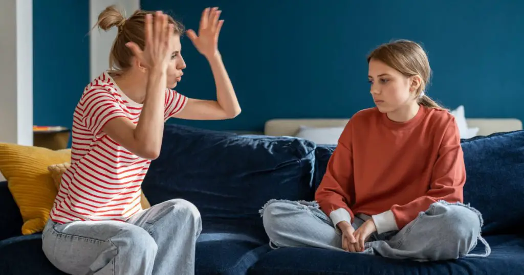 Young angry emotional mother screaming at troubled teen daughter, furious parent mom dealing with teenage behavior problems, yelling at upset adolescent girl while sitting together on sofa at home
