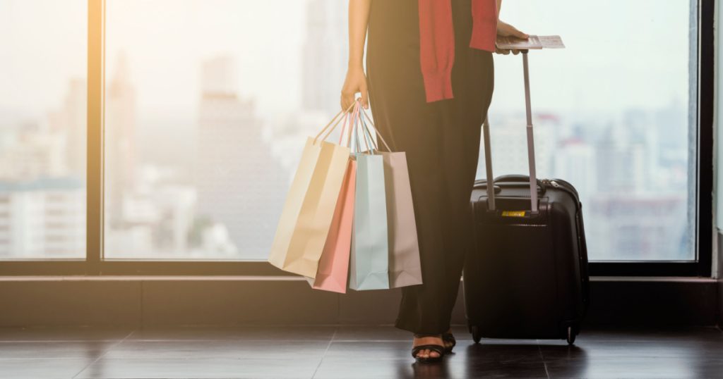 Advertising, Business, Travel, Shopping Concept - Cropped image of woman standing at the window in airport while carrying suitcase, shopping bags, passport and boarding pass. Copy space / Select focus
