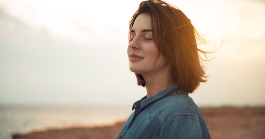 Great pleasure. Charming calm young woman is standing near sea with closed eyes and expressing delight. She is posing against wonderful sunset while enjoying last rays of the sun
