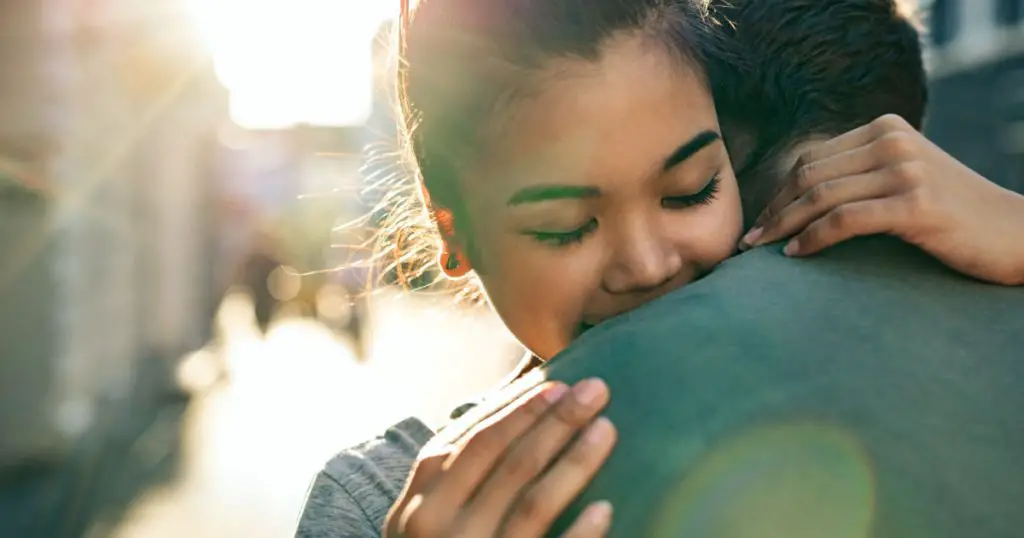 Young woman with her eyes closed hugging her boyfriend while standing together on a city street in the late afternoon
