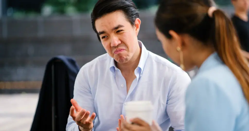 A handsome and young Asian business man in a crisp white shirt is talking to a woman in a suit. He is making a reluctant, grudging and unimpressed expression on his face as he thinks something over.
