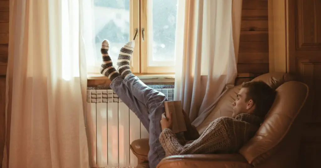 Young man in warm sweater reading book while relaxing on armchair by the window and electric radiator inside cozy log cabin
