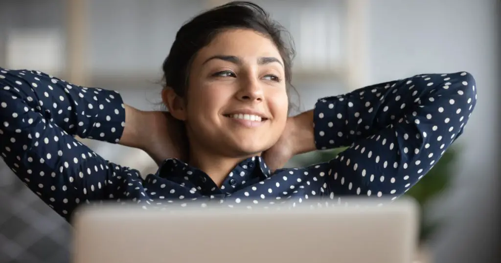 Happy satisfied indian woman rest at home office sit with laptop hold hands behind head, dreamy young lady relax finished work feel peace of mind look away dream think of future success concept
