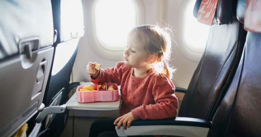 4 year old preschooler girl eating snacks from lunch box while travelling by plane
