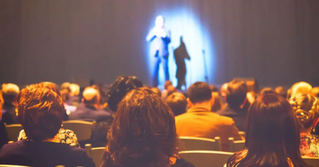 Man appears on stage in theater with a lot of people.
