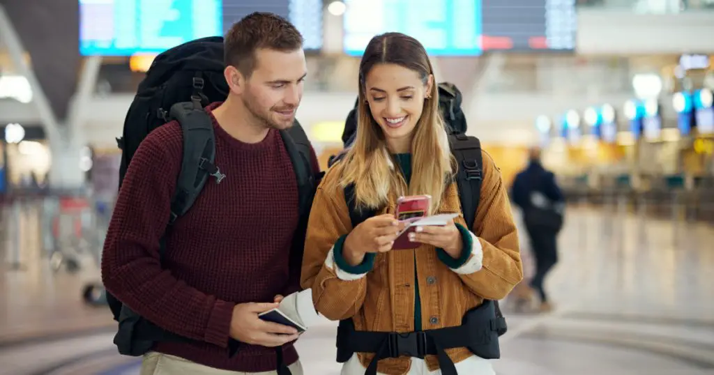 Happy couple, airport and phone with ticket, travel app and adventure with excited face, conversation and smile. Man, woman and smartphone for digital booking of hotel, taxi or bus for transportation

