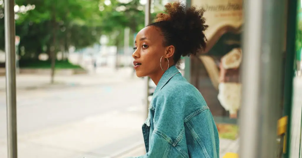 Cute girl with ponytail, wearing denim jacket, in crop top with national pattern, sitting at the bus stop and using her mobile phone.
