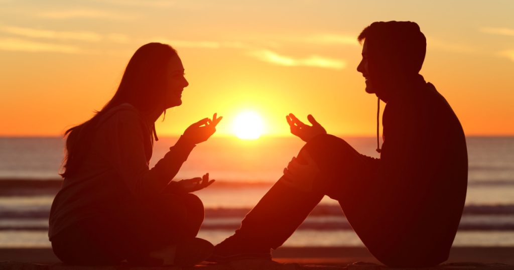 Side view of a full body of two friends or couple silhouette of teens sitting and talking at sunrise on the beach with the sun in the middle
