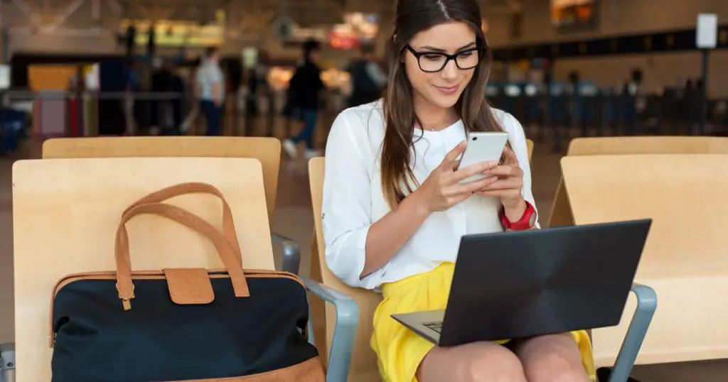 Airport Young female passenger on smart phone and laptop sitting in terminal hall while waiting for her flight. Air travel concept with young casual woman sitting with hand luggage suitcase.
