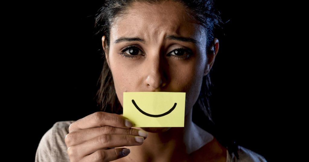 young beautiful Latin sad and depressed latin girl holding paper hiding her mouth behind a fake drawn smile pretending to be happy in depression concept isolated on black background
