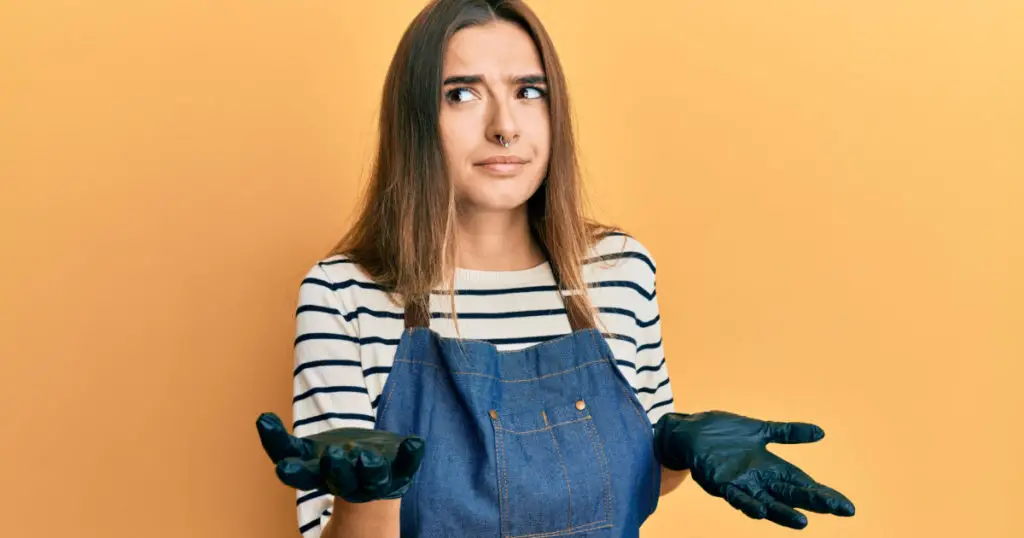Young hispanic woman wearing barber apron clueless and confused expression with arms and hands raised. doubt concept.