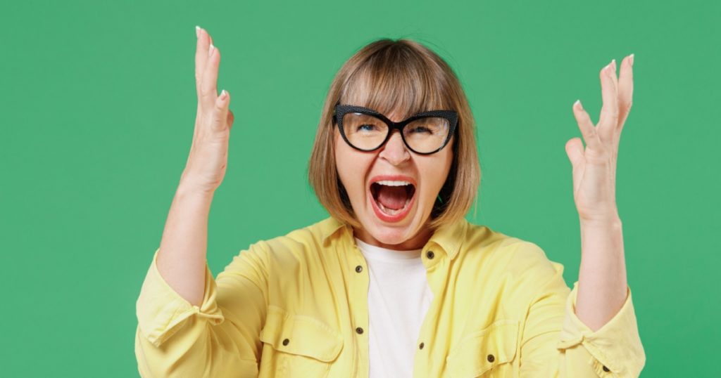 Elderly angry displeased irritated furious caucasian woman 50s in glasses yellow shirt spread hands loo camera scream shout isolated on plain green background studio portrait People lifestyle concept