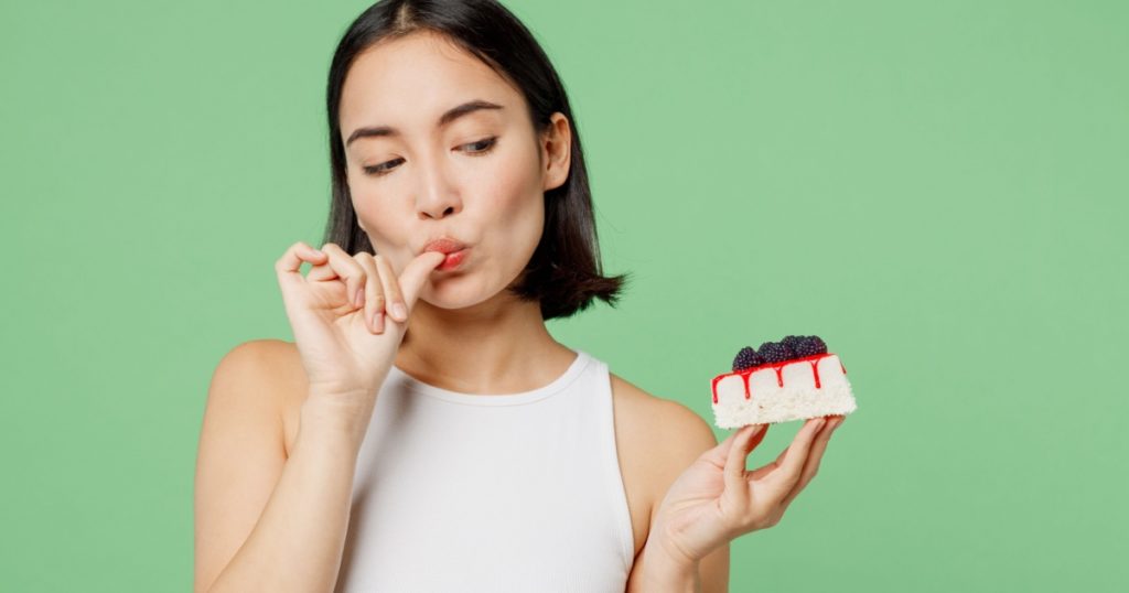 Young happy fun woman wear white clothes holding in hand pice of cake dessert lick fingers isolated on plain pastel light green background. Proper nutrition healthy fast food unhealthy choice concept