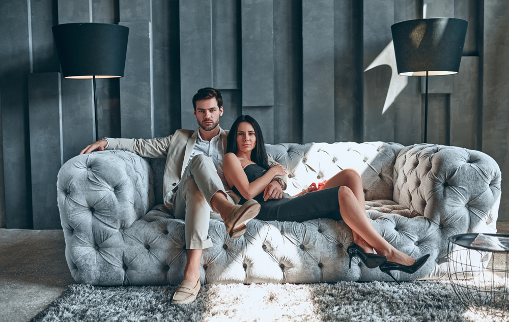 Stylish couple sitting on sofa together. Handsome bearded man in suit and attractive woman in black dress at home.