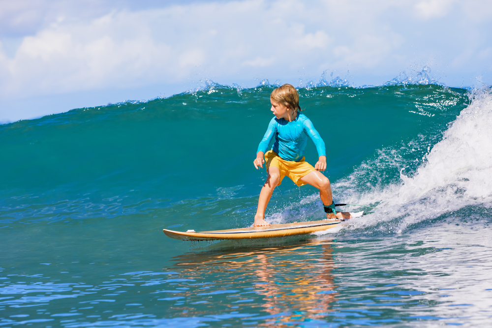 Happy surf boy - young surfer learn to ride on surfboard with fun on sea waves. Active family lifestyle, kids outdoor water sport lessons, swimming activity in surfing camp. Summer vacation with child
