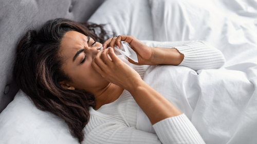 Sick black woman suffering from running stuffy nose and sore throat. Upset ill African American lady lying in bed, blowing her nose using paper napkin tissue, banner. Cold And Flu Concept