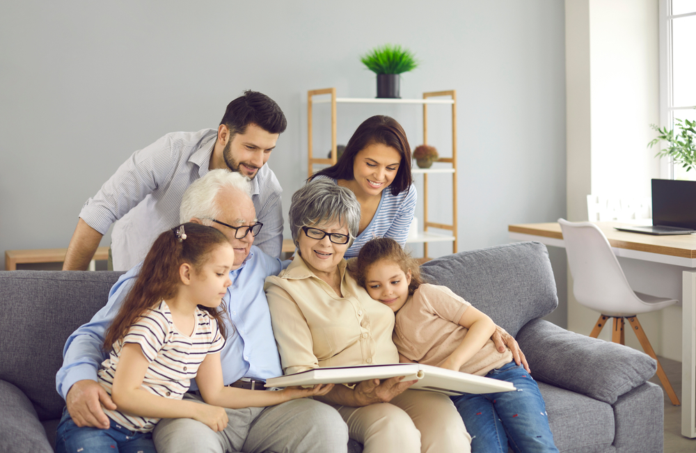 Happy extended family reading together. Young parents, little kids and mature grandparents sitting on sofa at home, enjoying book of good kind educational stories or looking through family photo album