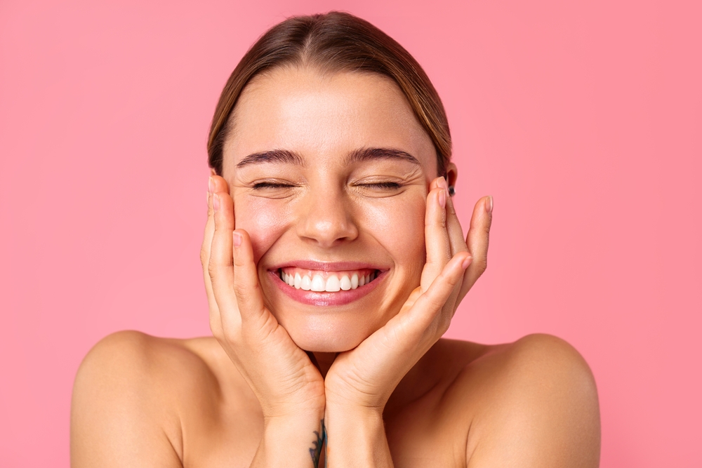 A cheerful young woman flashes a bright smile against a pink backdrop, showcasing her clear, radiant skin and natural beauty. Perfect for wellness, skincare, and positivity concepts.