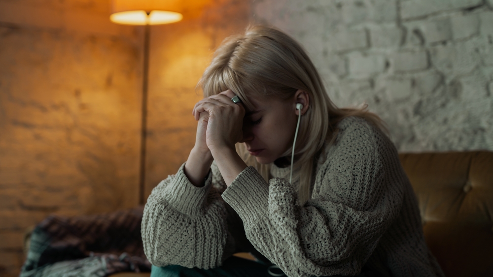 Sad Looking Young and Blonde Caucasian Woman Listening to Music from Her Ear Phones and Thinking Hard Over Life. Warm Light Coming From the Corner of The Room. White Brick Wall Living Room.