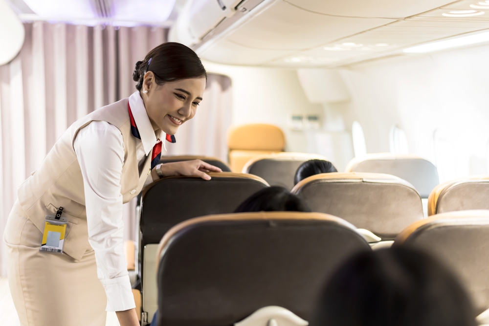 Friendly Asian female cabin crew smiling while talking to passenger on plane, happy air hostess woman working on board, cheerful Asian flight attendant woman taking care of treaveller during flight