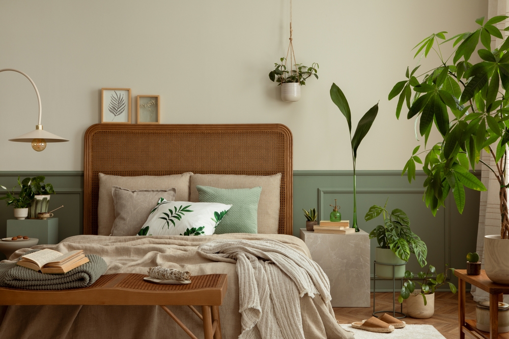 Warm and cozy bedroom interior with mock up poster frame, boho bed, beige bedding, green wall with stucco, books, brown slippers, plants in pots and personal accessories. Home decor. Template.